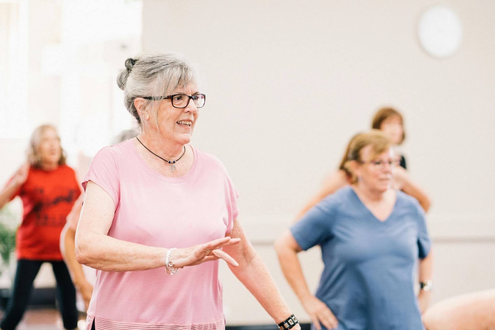 A senior woman in a pink shirt and glasses smiles while participating in a group exercise class.