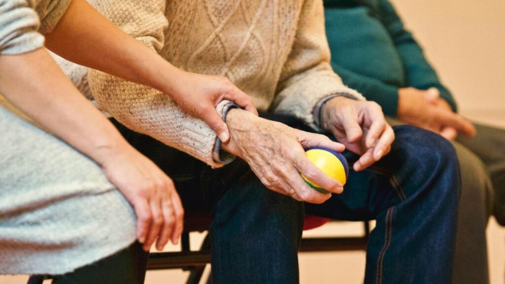 Caregiver supporting an older adult's arm during a seated activity with a small therapy ball.