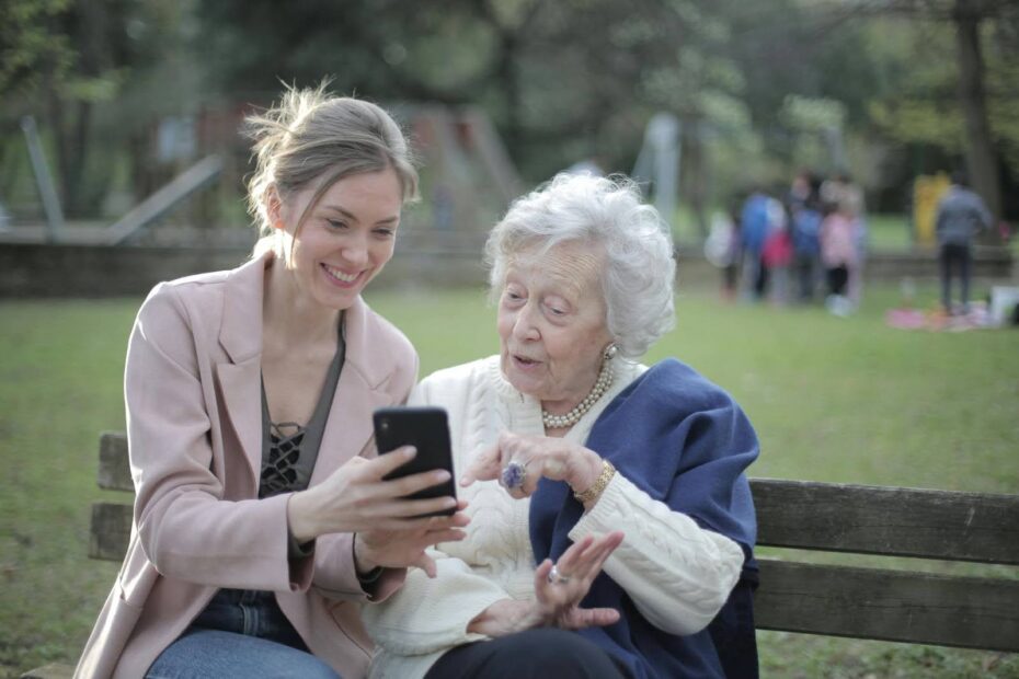 Adult daughter and elderly mother smiling together while looking at a smartphone on a park bench.